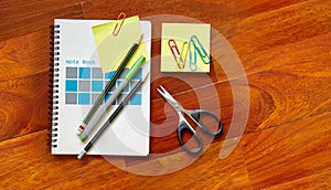 Notebook with yellow sticky note clipped on the cover page, pencils, scissor on the parquet wooden floor with copy space or text s