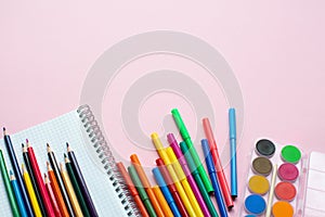 Notepad with color pencils and different school supplies on pink background with copy space.