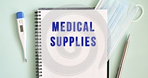 Notepad, medical mask, thermometer and pen on a blue background. MEDICAL SUPPLIES text in a notebook.