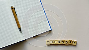 Notepad with pen and wooden word `Thursday` on a beige paper background. Top view. Close-up.