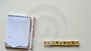 Notepad with pen and wooden word `Wednesday` on a beige paper background. Top view. Close-up.