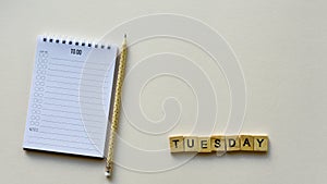 Notepad with pencil and wooden word `Tuesday` on a beige paper background. Top view. Close-up.