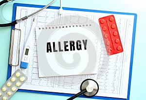 A notepad with the text ALLERGY lies on a medical clipboard with a stethoscope and pills on a blue background.