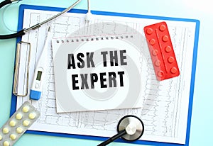 A notepad with the text ASK THE EXPERT lies on a medical clipboard with a stethoscope and pills on a blue background.