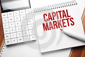 Notepad with text Capital Markets on wooden background with calculator and red marker