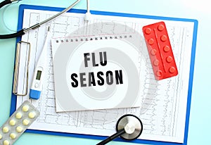 A notepad with the text FLU SEASON lies on a medical clipboard with a stethoscope and pills on a blue background.