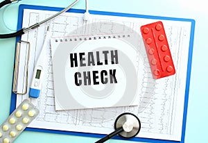 A notepad with the text HEALTH CHECK lies on a medical clipboard with a stethoscope and pills on a blue background.