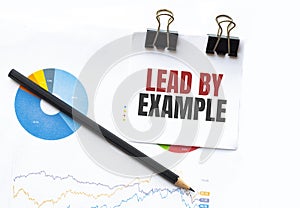 Notepad with text LEAD BY EXAMPLE on business charts and pen