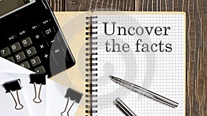 Notepad with text Uncover The Facts on the wooden background with clips, pen and calculator