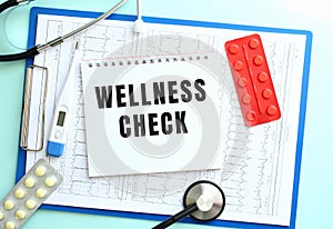 A notepad with the text WELLNESS CHECK lies on a medical clipboard with a stethoscope and pills on a blue background.