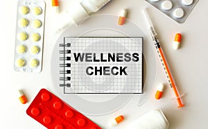 Notepad with text WELLNESS CHECK on a white background. Nearby are various medicines.