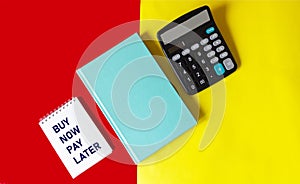 Notepad with the words BUY NOW PAY LATER on a red and yellow background with a green diary and a calculator