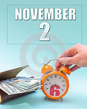 November 2nd. Hand holding an orange alarm clock, a wallet with cash and a calendar date. Day 2 of month.