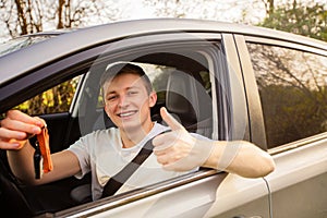 Novice driver holding car keys out of the window, showing thumb up positive gesture and smiling. New driver on the road concept