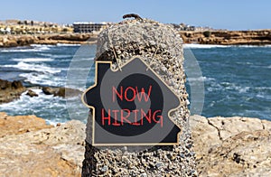 Now hiring symbol. Concept words Now hiring on beautiful black chalk blackboard. Beautiful stone sea blue sky background. Business