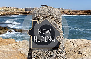 Now hiring symbol. Concept words Now hiring on beautiful black chalk blackboard. Beautiful stone sea blue sky background. Business