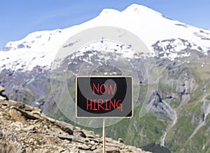 Now hiring symbol. Concept words Now hiring on beautiful black chalk blackboard. Beautiful mountain Elbrus blue sky background.