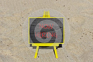 Now hiring symbol. Concept words Now hiring on beautiful black chalk blackboard. Beautiful sea sand beach background. Business