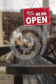 Now - We Are Open sign in front of blurred people outside a cafe enjoying diner, drinks and coffee.