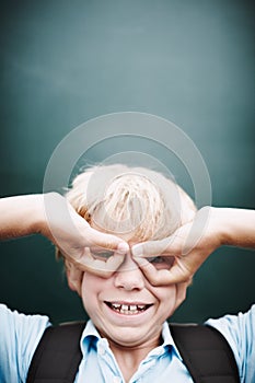 Now you see me. A funny young boy making glasses with his hands against a classroom blackboard.