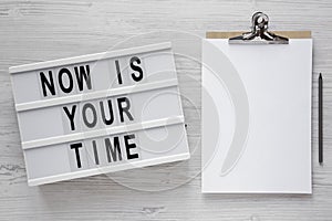 `Now is your time` words on a lightbox, clipboard with blank sheet of paper on a white wooden surface, top view. Overhead, from