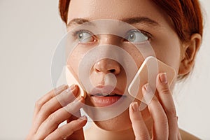 Nude makeup. Beautiful young girl applying face foundation over white studio background. Well-kept skin