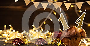 Number 14 gold burning candle in a cupcake against celebration wooden background with lights. Birthday cupcake. Copy