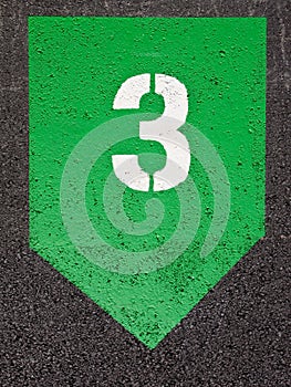 Number three stencilled in white paint on a green geometric symbol on asphalt surface