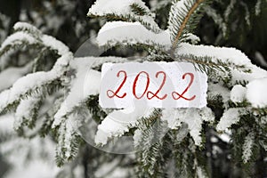 The numbers 2022 are written on paper with a snowy Christmas tree in the background. Snow is falling. Selective focus