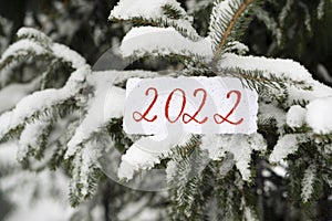 The numbers 2022 are written on paper with a snowy Christmas tree in the background. Snow is falling. Selective focus, blurred