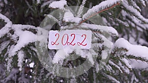 The numbers 2022 are written on paper with a snowy Christmas tree in the background. Snow is falling. Selective focus, blurred win