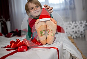 numbers 2023 are written on boy's bare feet, child wears a little Santa hat on his toes