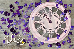 2020 numbers on a wooden background next to purple pansies flowers and a pink clock showing five minutes to midnight