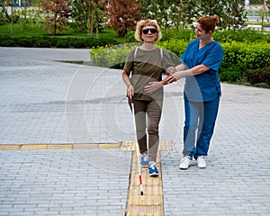 A nurse accompanies an elderly blind woman on a walk.