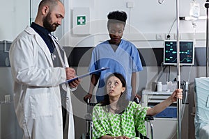 Nurse attending woman with illness sitting in wheelchair holding intravenous pole while doctor holding clipboard
