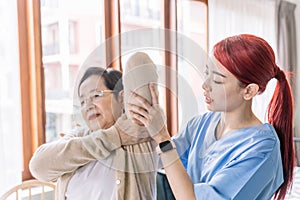 Nurse caregiver wearing scrubs exercises with a senior Asian woman to relieve a frozen shoulder of the senior patient.