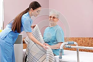 Nurse covering senior woman with plaid on bed in hospital ward