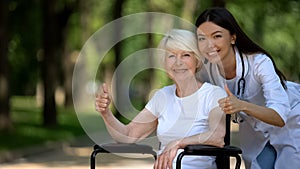 Nurse and elderly woman in wheelchair smiling at camera and showing thumbs-up photo