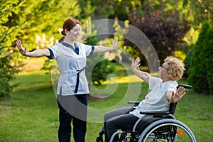 A nurse is exercising with an elderly patient in a wheelchair outdoors.