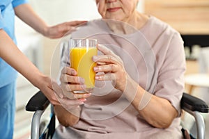 Nurse giving glass of juice to elderly woman indoors. Assisting senior people