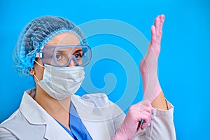 Nurse in goggles puts on protective gloves for work, blue background. A doctor in a white coat and protective equipment for the