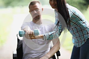 Nurse helping disabled young man in wheelchair to lift dumbbell closeup