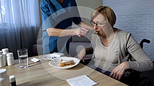 Nurse helping sick old female to eat, medical support and care, sadness