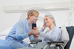 Nurse helps with cell phone to contact the elderly lady`s family in the wheelchair near bed in hospital room, concept of