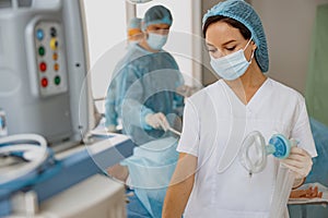 Nurse holding breathing mask for anesthesia standing in operation room with colleagues on background