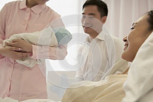 Nurse holding newborn baby in the hospital with mother lying on the bed and the father beside her