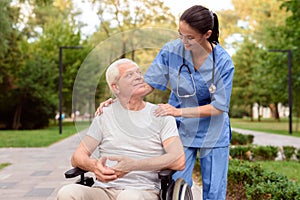 The nurse looks carefully at the old man, who is sitting in a wheelchair and smiling at him