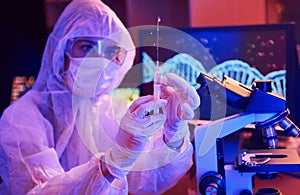 Nurse in mask and white uniform, holding syringe and sitting in neon lighted laboratory with computer and medical