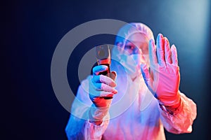 Nurse in mask and white uniform standing in neon lighted room and holding infrared thermometer. Stop coronavirus