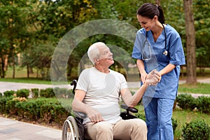 A nurse measures the pulse of a smiling old man who sits in a wheelchair in a green park